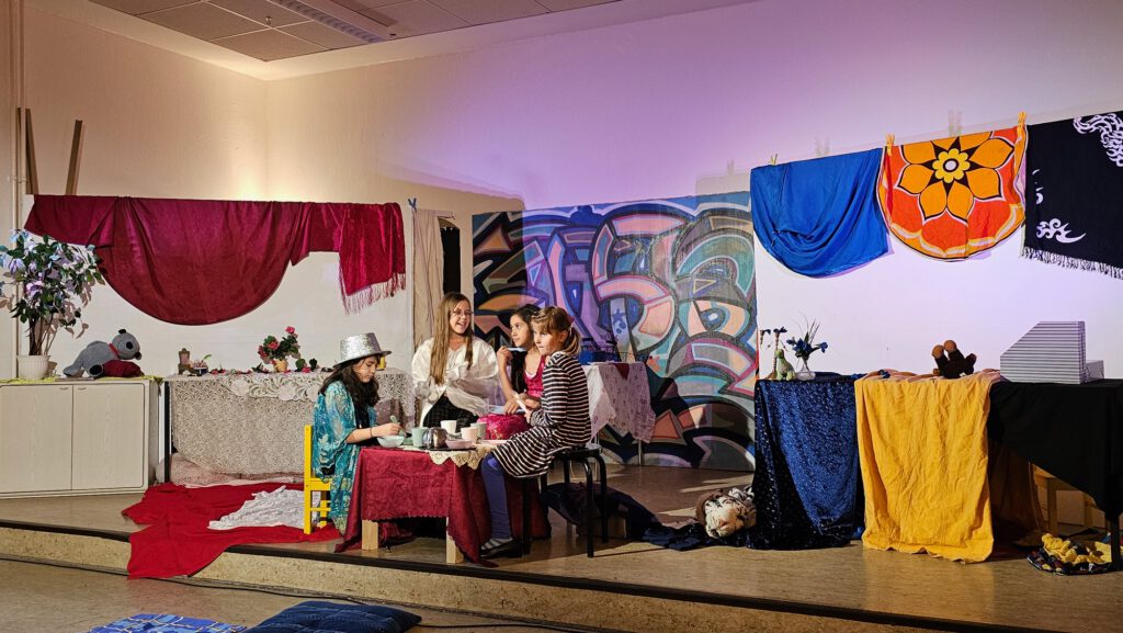 Drei Kinder sitzen an einem Tisch auf einer Bühne, der Hintergrund ist bunt.